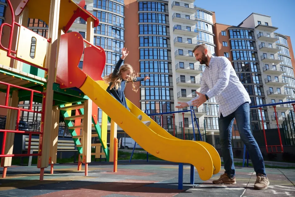 Man reaching for young girl sliding down playground slide.
